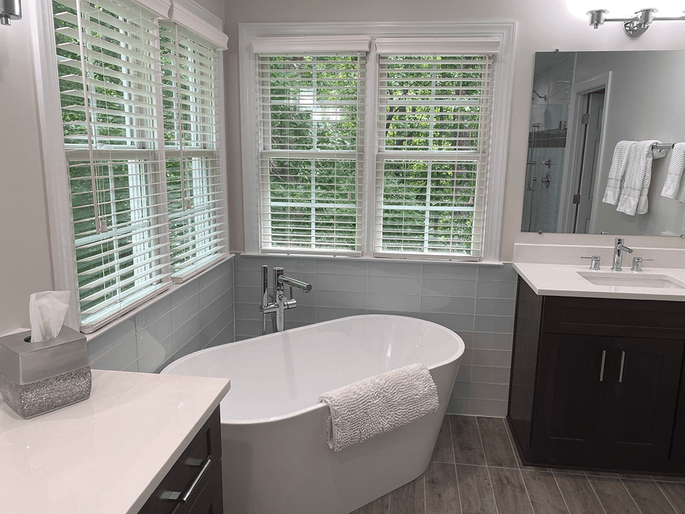A large, white, stand alone bath tub in the corner of a bathroom with light gray tiled walls and vanities with dark cabinets and light countertops.