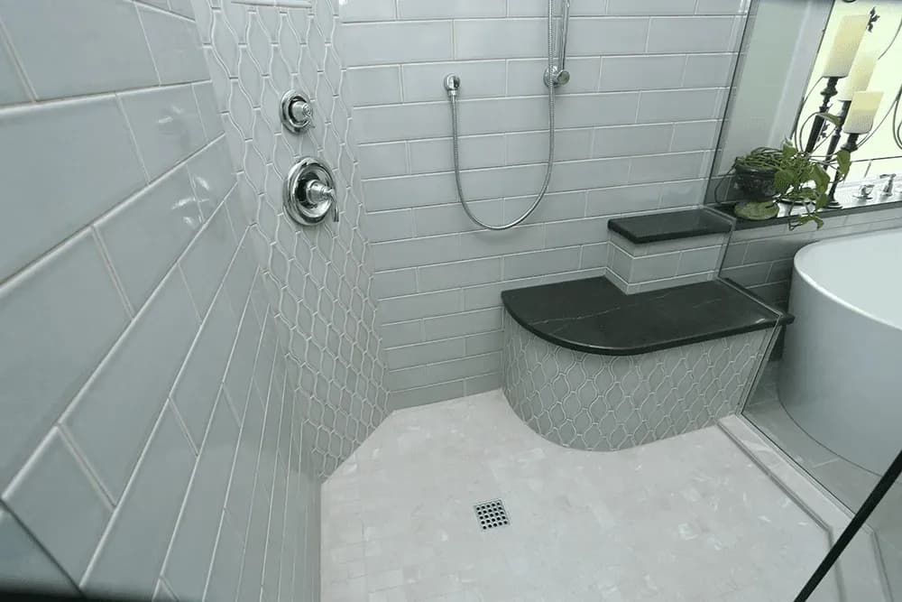 A shower with gray mixed geometric tiled walls and built-in bench seat with dark stone top.