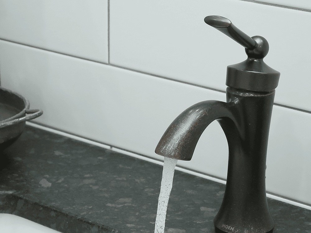 A close-up of a running, oil-rubbed bronze faucet in a bathroom with white tile walls and dark countertop.